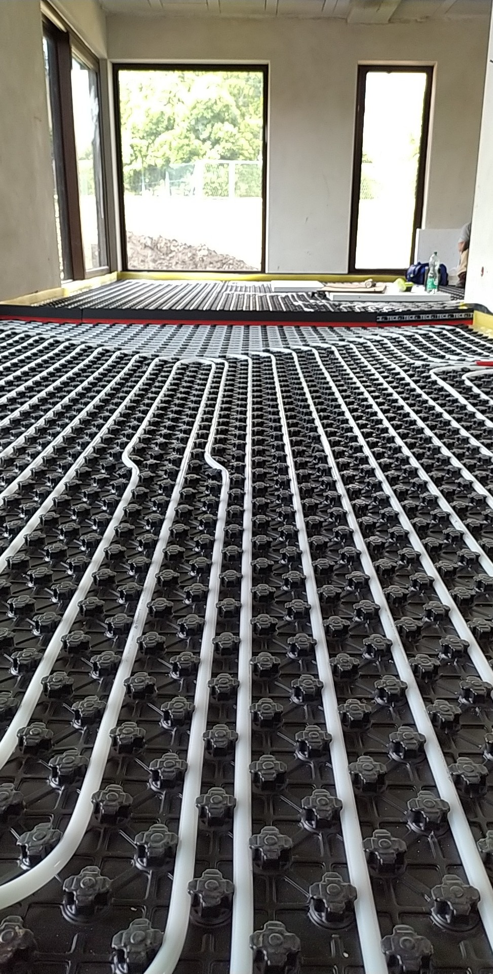 Ułożona instalacja ogrzewania podłogowego w surowym wnętrzu z dużymi oknami. Białe rurki na czarnej macie, widok z perspektywy podłogi.