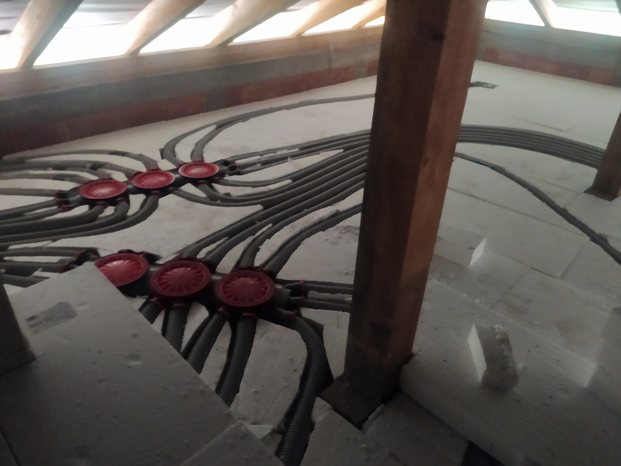 Szare rury instalacji elektrycznej w peszlu, rozchodzące się promieniście od czerwonych puszek podłączeniowych w surowym stanie poddasza. Widoczne drewniane elementy konstrukcji.