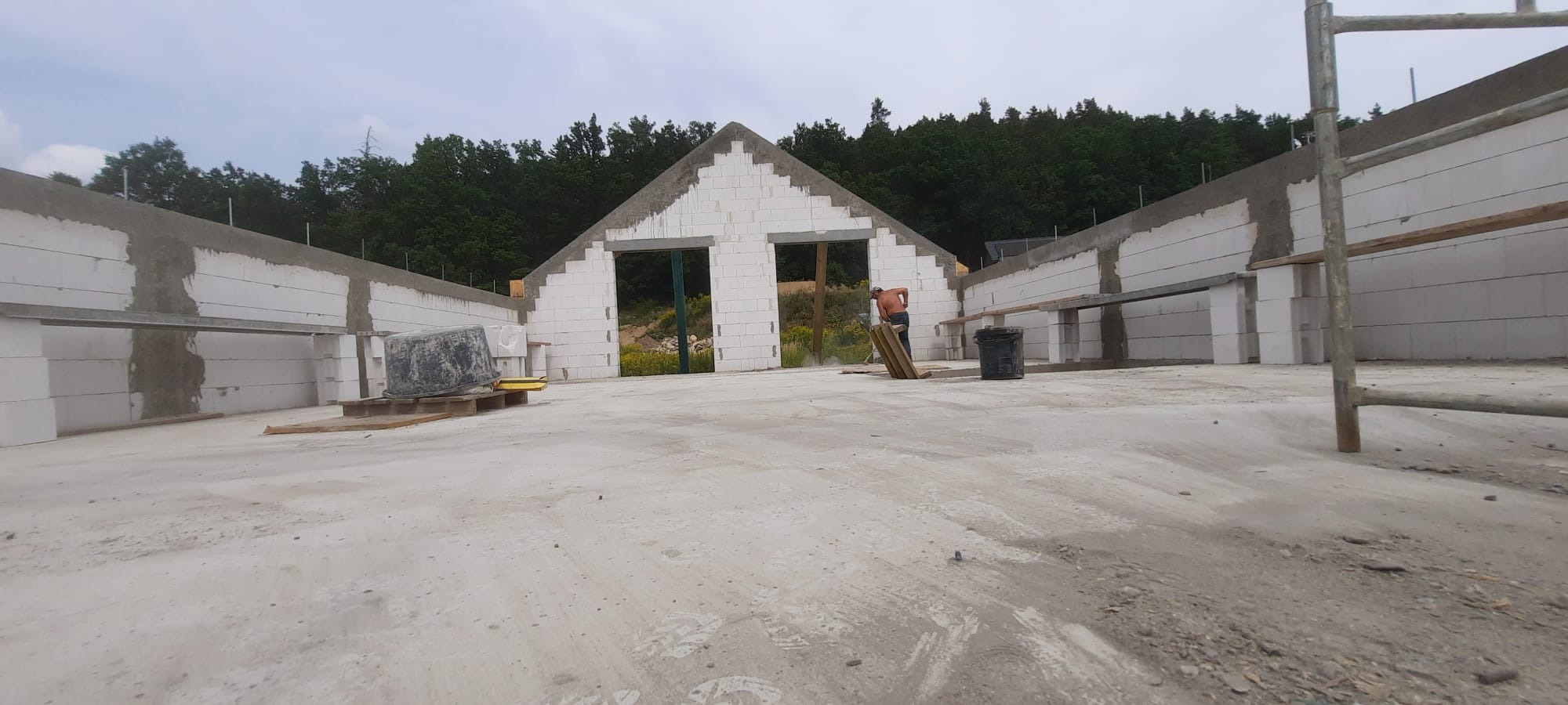 Wznoszenie ścian budynku z białych bloczków, widoczne otwory okienne i drzwiowe. Prace murarskie w toku, widoczne rusztowania i narzędzia. Na drugim planie zieleń drzew.