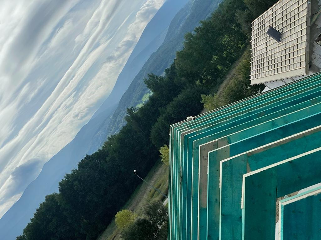 Szkielet dachu w trakcie budowy, z zielonymi belkami na tle gór i nieba. Widoczne elementy konstrukcyjne i fragment gotowego budynku.