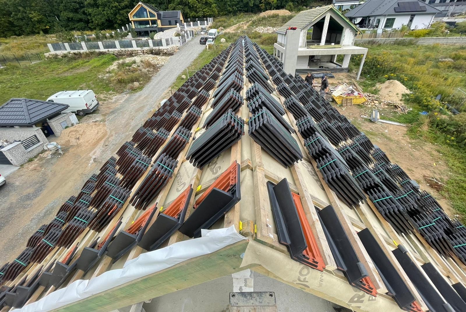 Wysoki kąt na dach w trakcie krycia dachówką ceramiczną. Widoczne przygotowane rzędy dachówek z elementami mocującymi. W tle zabudowa jednorodzinna i zieleń.