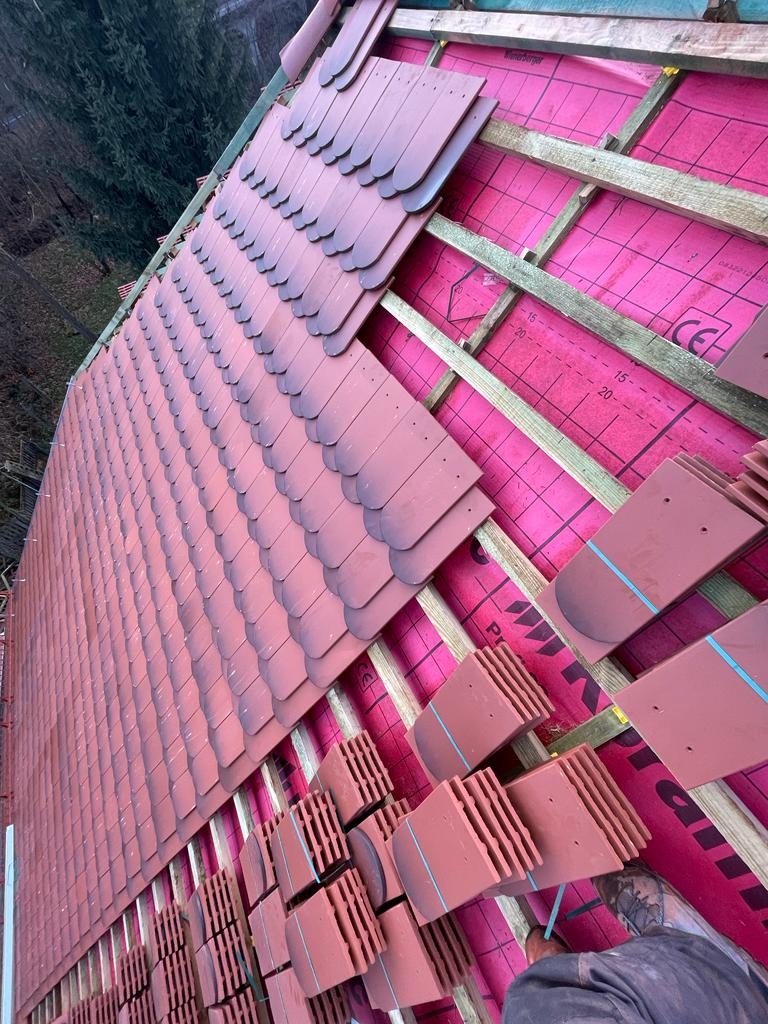 Układanie dachówki karpiówki na różowej membranie, widoczne drewniane łaty i fragmenty dachówek w pakietach. Praca w toku, widok z dołu na dach.