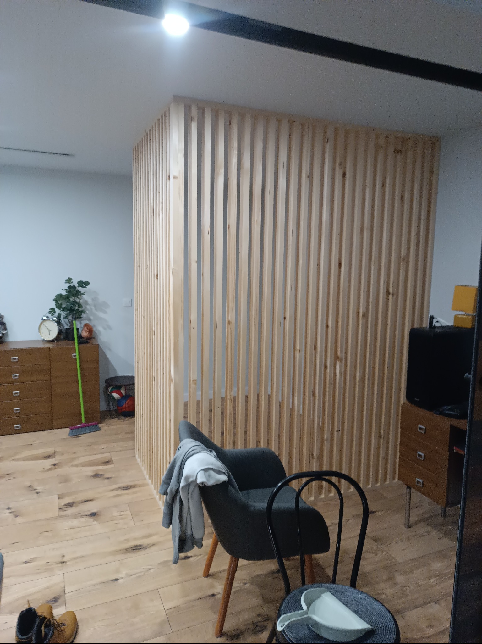 Drewniana ścianka działowa z pionowych listew oddziela przestrzeń w pokoju. Na pierwszym planie krzesła, w tle komoda i podłoga z paneli.