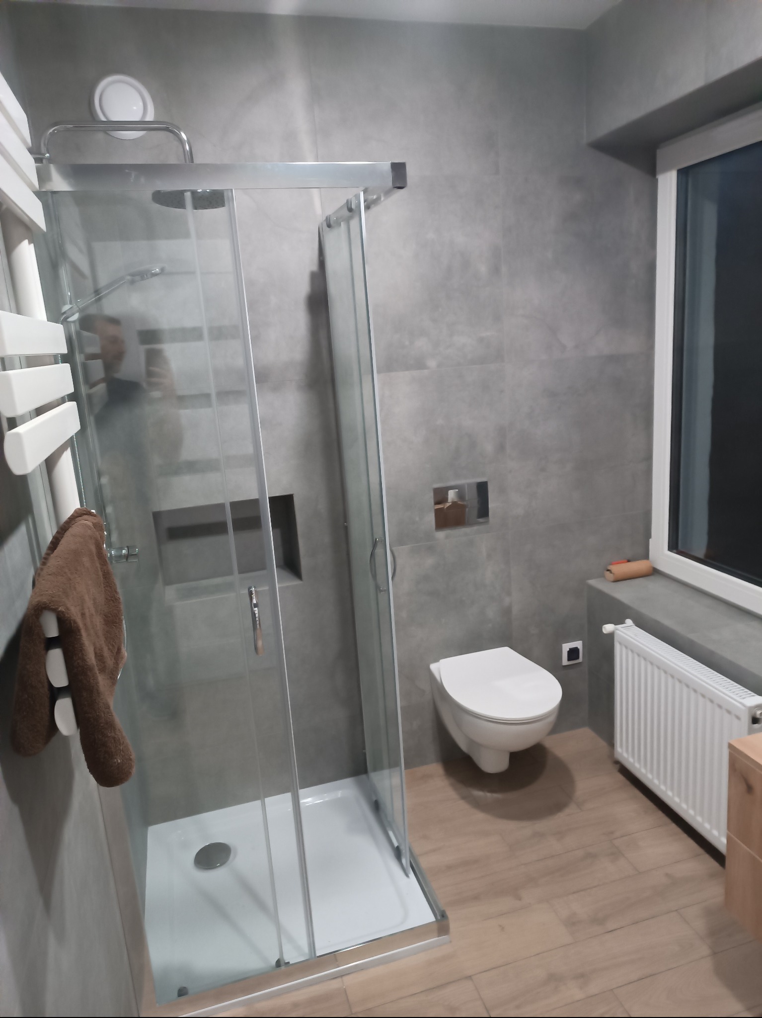 Nowoczesna łazienka po remoncie: szara glazura, kabina prysznicowa ze srebrnymi ramami, podwieszana toaleta, drewnopodobne panele i biały grzejnik.