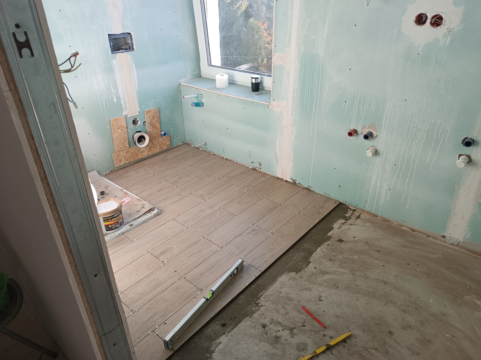 Wnętrze łazienki w trakcie remontu: częściowo ułożone płytki podłogowe o wzorze drewna, ściany z płyt gipsowo-kartonowych, poziomica, puszki elektryczne.