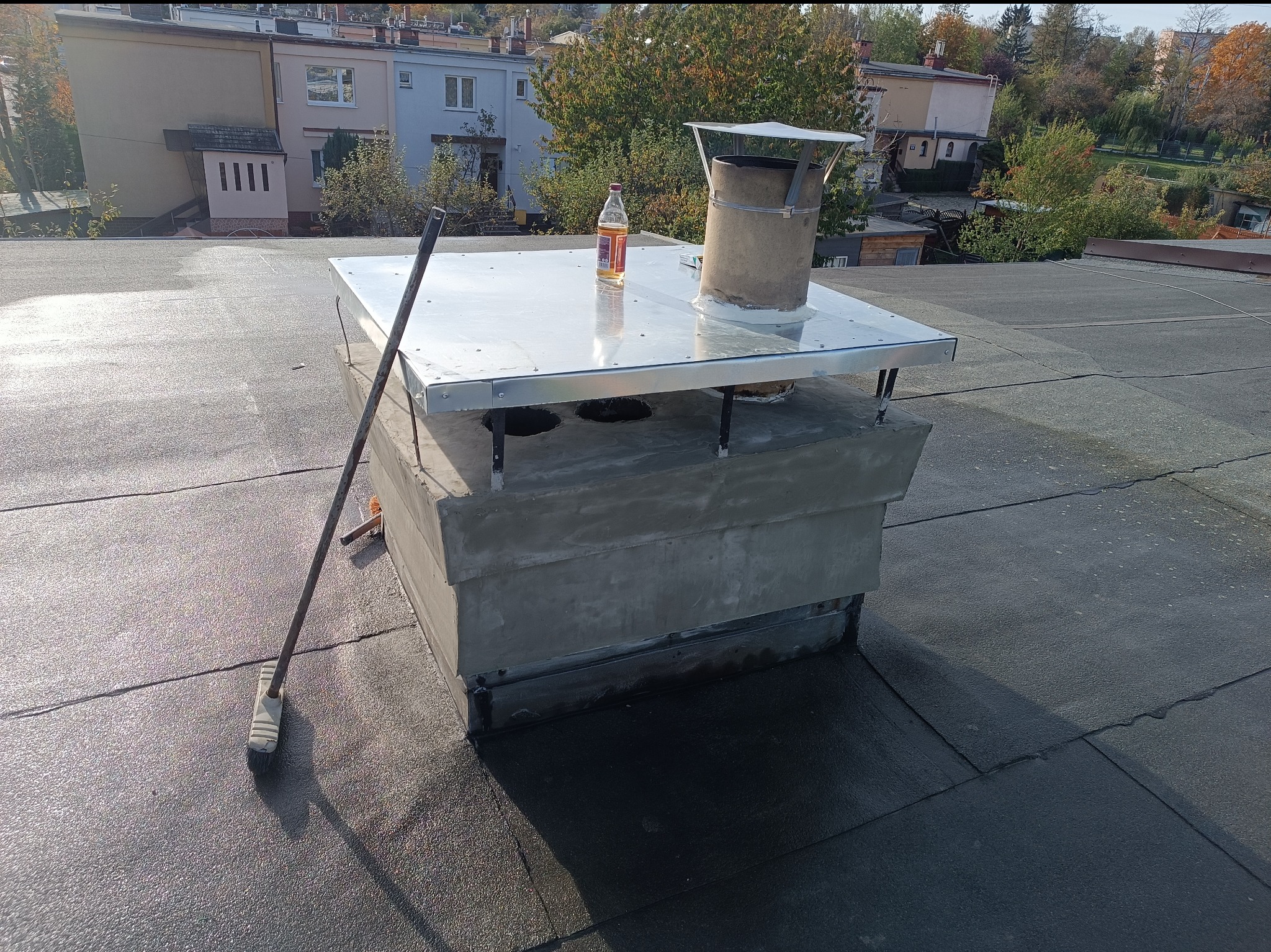 Błyszczący, metalowy komin z osłoną na płaskim dachu, obok butelka i szczotka. W tle budynki mieszkalne i drzewa.