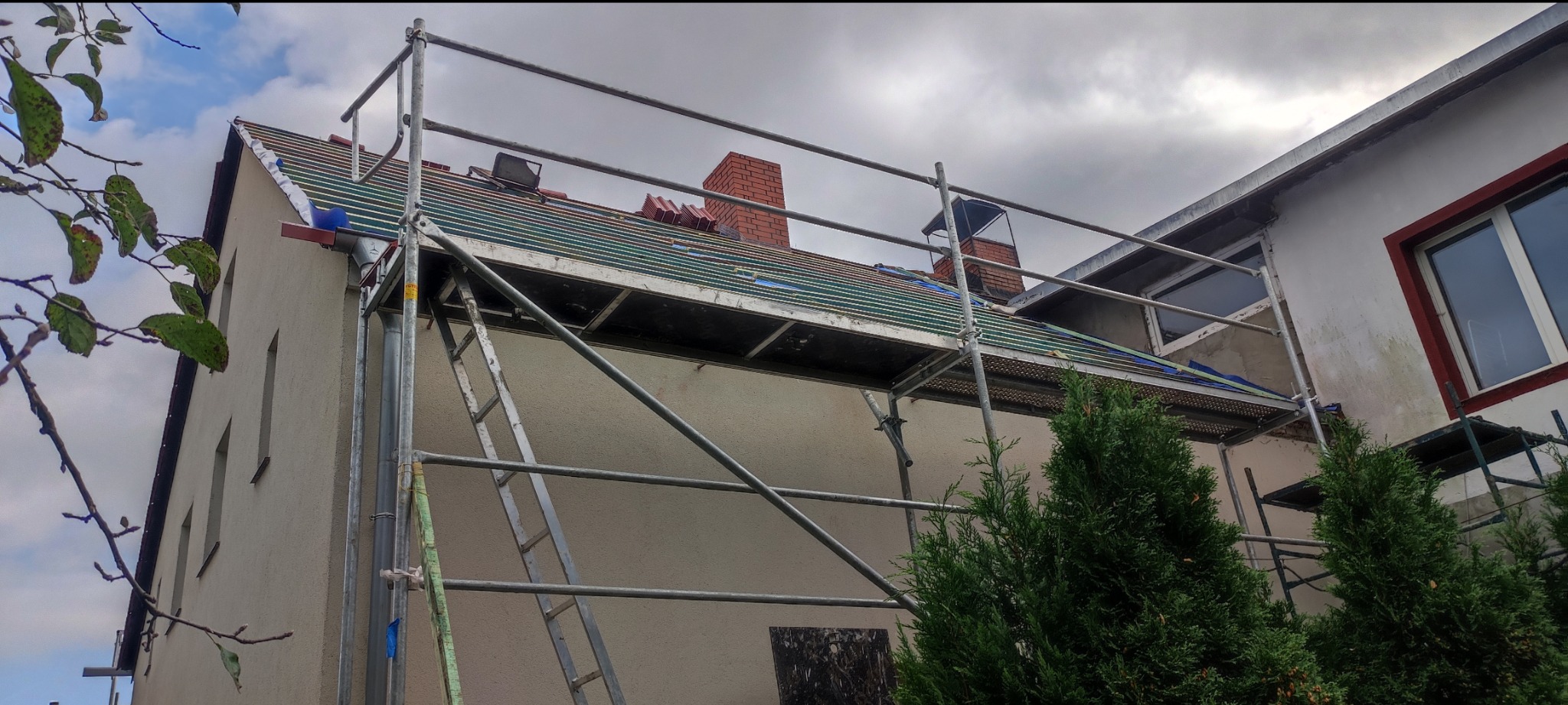 Dom w trakcie remontu dachu z użyciem rusztowania. Widoczne nowe elementy konstrukcyjne i komin. Częściowo zdjęte dachówki. Prace dekarskie w toku.