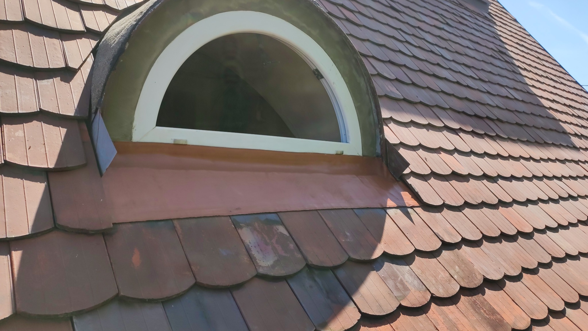 Dach z brązowej dachówki ceramicznej z oknem w kształcie półkola, biała rama okienna, widoczne obróbki blacharskie. Dachówka karpiówka.