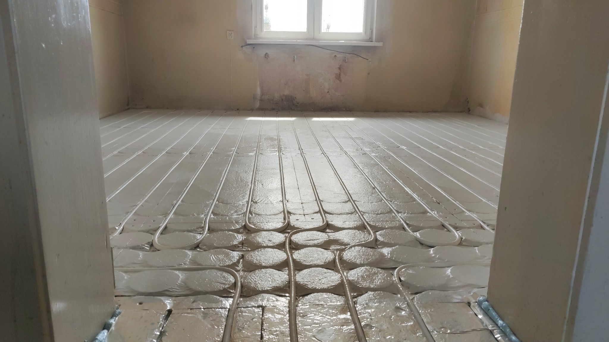 Biała instalacja ogrzewania podłogowego, z widocznymi rurami i folią izolacyjną, rozłożona w pustym pomieszczeniu, gotowa do zalania posadzką. Widok zza framugi drzwi.