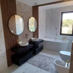 Nowoczesna łazienka z marmurowymi płytkami, dwiema umywalkami nablatowymi na czarnych szafkach, owalnymi lustrami z podświetleniem, wolnostojącą wanną i bidetem z złotą armaturą, oraz dekoracyjnymi...