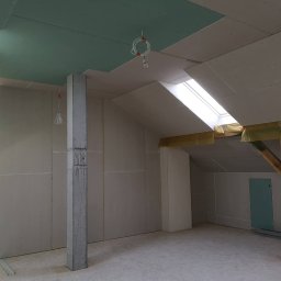 Usługi budowlane - Poddasze w trakcie adaptacji: ściany i sufit wyłożone płytami gipsowo-kartonowymi, widoczne fragmenty drewnianej konstrukcji dachu zabezpieczone folią, surowy betonowy filar z zaciekami, tymczasowe...