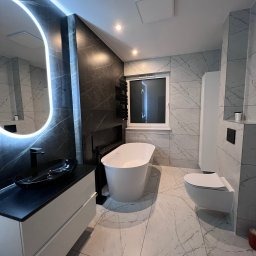 Nowoczesna łazienka z marmurowymi płytkami, owalnym lustrem z podświetleniem LED, wolnostojącą białą wanną i czarną umywalką na białej szafce. Widoczne okno i grzejnik drabinkowy.