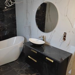 Nowoczesna łazienka z czarno-białymi marmurowymi płytkami, owalnym lustrem, białą wanną wolnostojącą i czarną szafką z owalną umywalką. Widoczne wystające kable elektryczne sugerują prace w toku.