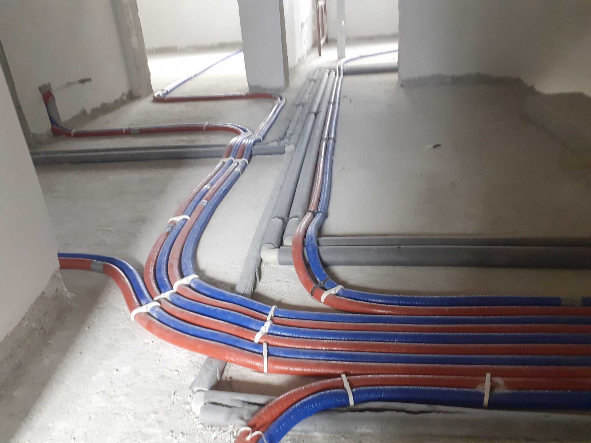 Rozprowadzone na podłodze rury instalacji hydraulicznej w kolorach czerwonym i niebieskim, połączone białymi opaskami zaciskowymi, w surowym stanie wykończenia wnętrza.