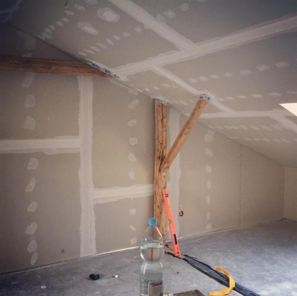 Poddasze w trakcie prac wykończeniowych: płyty gipsowo-kartonowe na skośnym suficie i ścianach, widoczne łączenia płyt, drewniana belka wzmacniająca konstrukcję, poziomica oparta o belkę, butelka...
