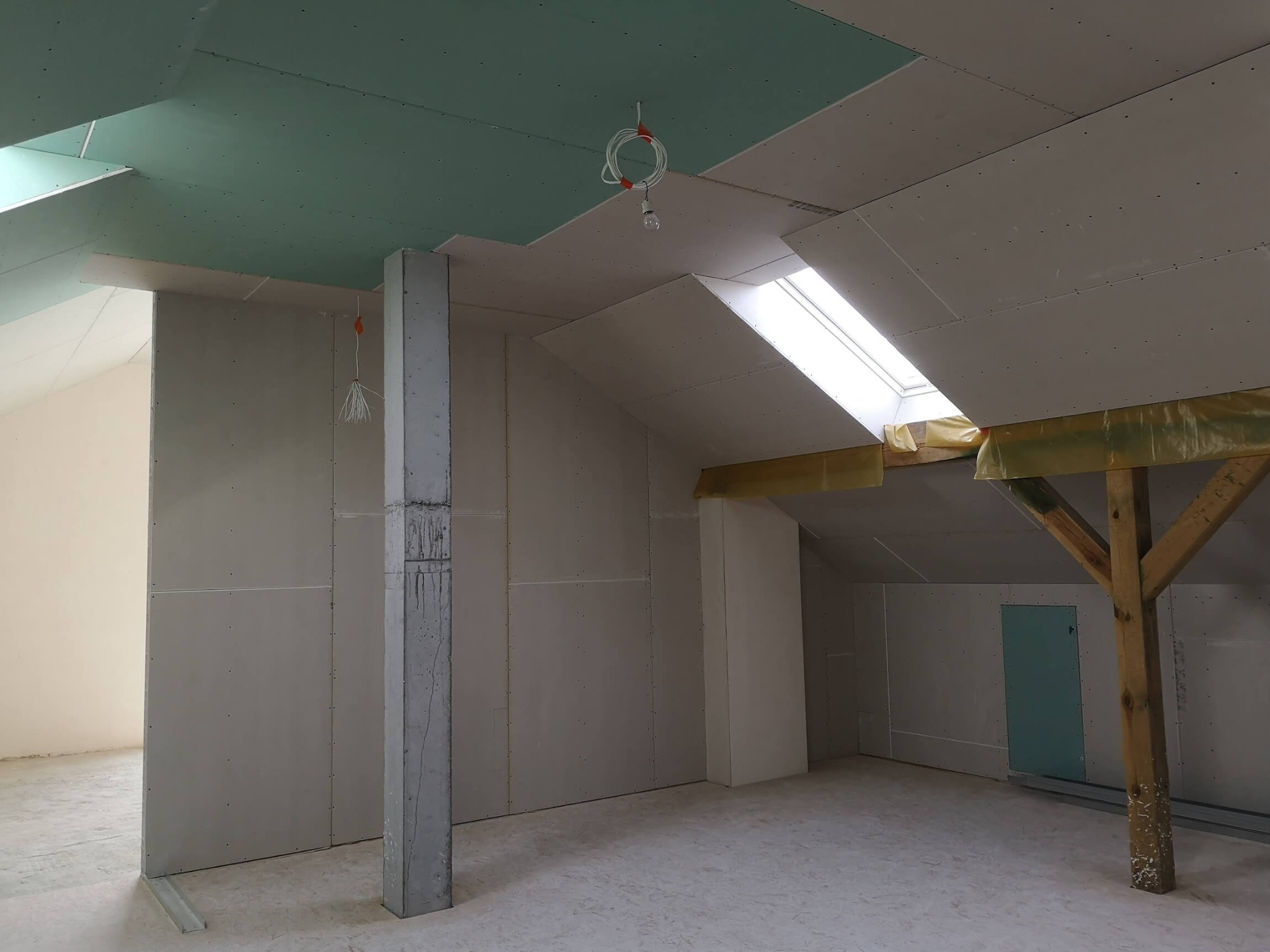 Poddasze w trakcie adaptacji: ściany i sufit wyłożone płytami gipsowo-kartonowymi, widoczne fragmenty drewnianej konstrukcji dachu zabezpieczone folią, surowy betonowy filar z zaciekami, tymczasowe...