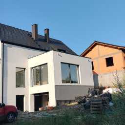 Usługi Ogólnobudowlane - Nowoczesny dom jednorodzinny z białymi ścianami i ciemną dachówką w trakcie prac wykończeniowych; widoczny samochód dostawczy i materiały budowlane.