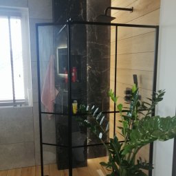 Usługi Ogólnobudowlane - Nowoczesna kabina prysznicowa z czarnymi profilami, szklanymi ściankami, deszczownicą i rośliną w doniczce, na tle płytek imitujących drewno i marmur.
