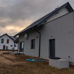 Usługi Ogólnobudowlane - Nowoczesny dom jednorodzinny z szarą elewacją i ciemną stolarką okienną, widok z boku, na pierwszym planie fragment gruntu z niebieskim wężem ogrodowym.