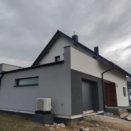 Usługi Ogólnobudowlane - Dwukondygnacyjny dom jednorodzinny z elewacją w dwóch odcieniach szarości i bieli, widoczny klimatyzator na szarej ścianie bocznej, teren wokół domu w trakcie prac wykończeniowych.