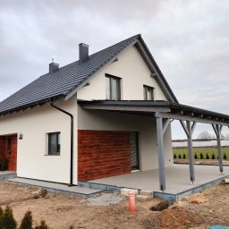 Usługi Ogólnobudowlane - Nowoczesny dom jednorodzinny z białymi ścianami, drewnianą elewacją fragmentu i szarą dachówką, zadaszony taras z drewnianą konstrukcją, widok zewnętrzny w pochmurny dzień.