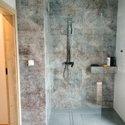 Usługi Ogólnobudowlane - Wykończona łazienka z kabiną prysznicową typu walk-in, wyłożoną płytkami w odcieniach szarości z delikatnym wzorem, z zamontowaną deszczownicą i półką na kosmetyki, widoczny odpływ liniowy...