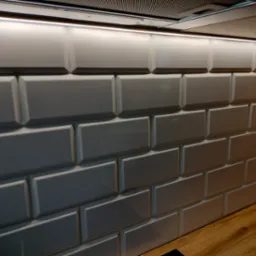 Ściana wyłożona białymi, błyszczącymi kafelkami typu metro w kuchni, oświetlona listwą LED pod szafkami, z widocznym fragmentem drewnianego blatu.