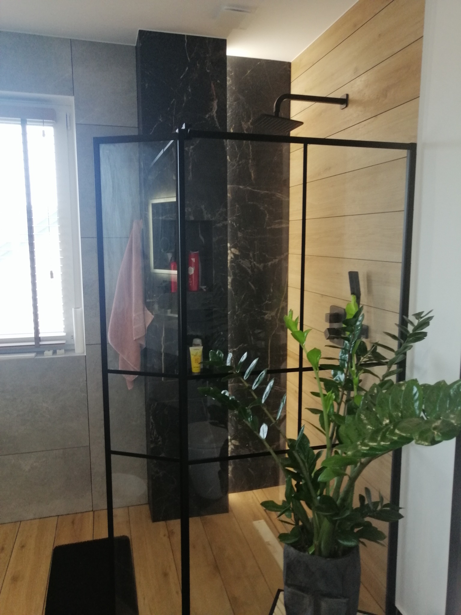 Nowoczesna kabina prysznicowa z czarnymi profilami, szklanymi ściankami, deszczownicą i rośliną w doniczce, na tle płytek imitujących drewno i marmur.