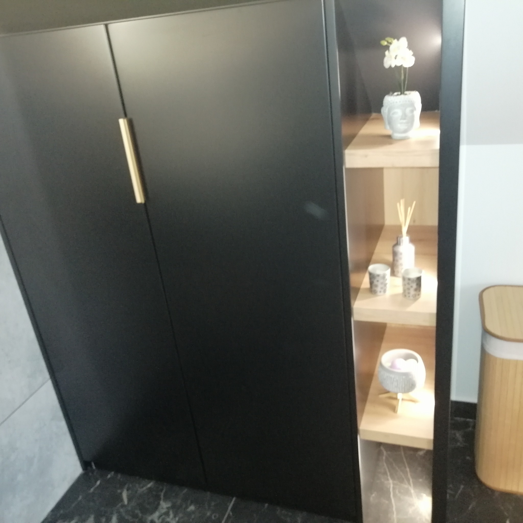 Czarny, matowy regał z zamkniętymi drzwiczkami z lewej strony i otwartymi półkami z prawej, na których stoją dekoracje w łazience, takie jak kwiaty w wazonie w kształcie głowy Buddy, dyfuzor...