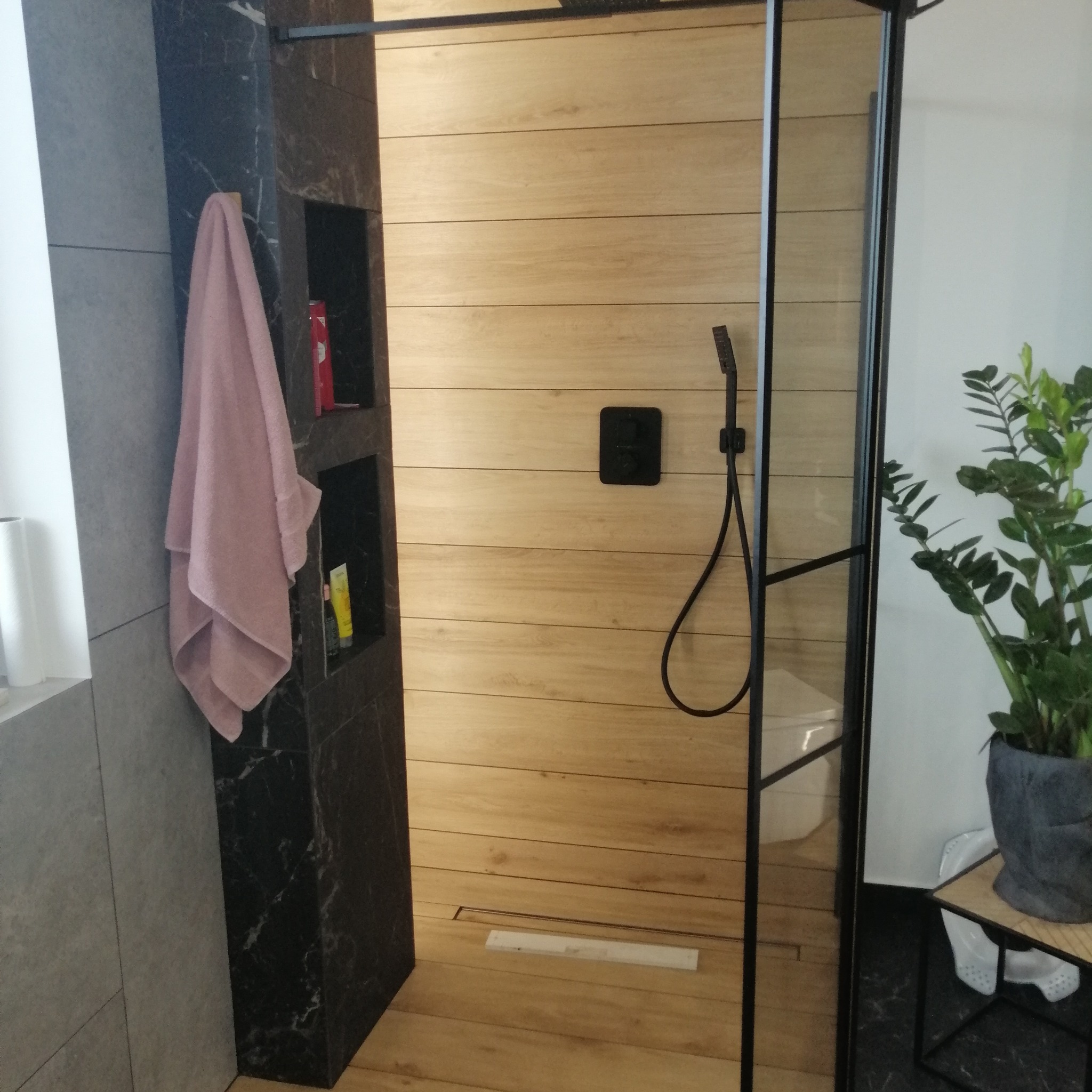 Nowoczesna kabina prysznicowa z drewnopodobnymi płytkami, czarną armaturą i marmurowym wykończeniem, z widocznym odpływem liniowym i rośliną doniczkową obok.