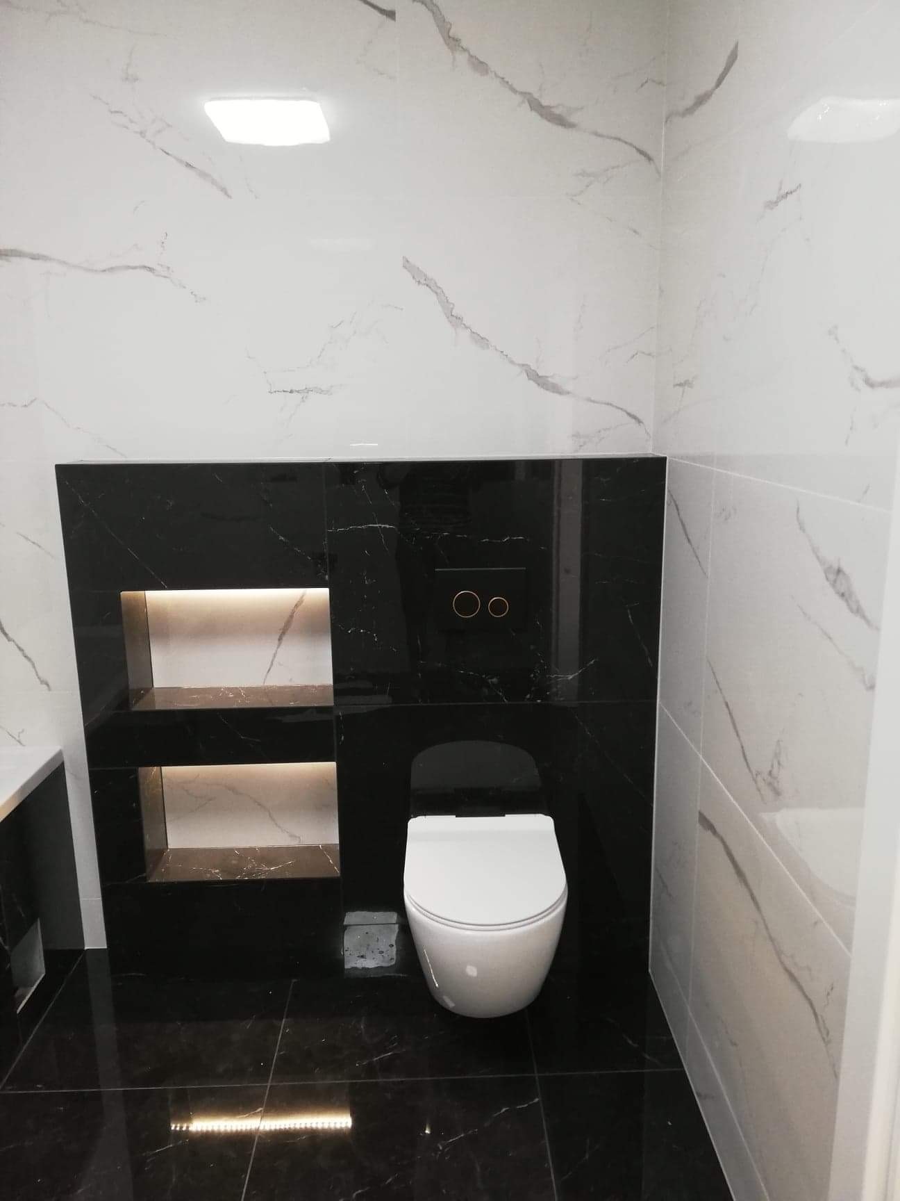 Nowoczesna łazienka z czarno-białymi płytkami imitującymi marmur, podwieszana toaleta i podświetlane wnęki w zabudowie.