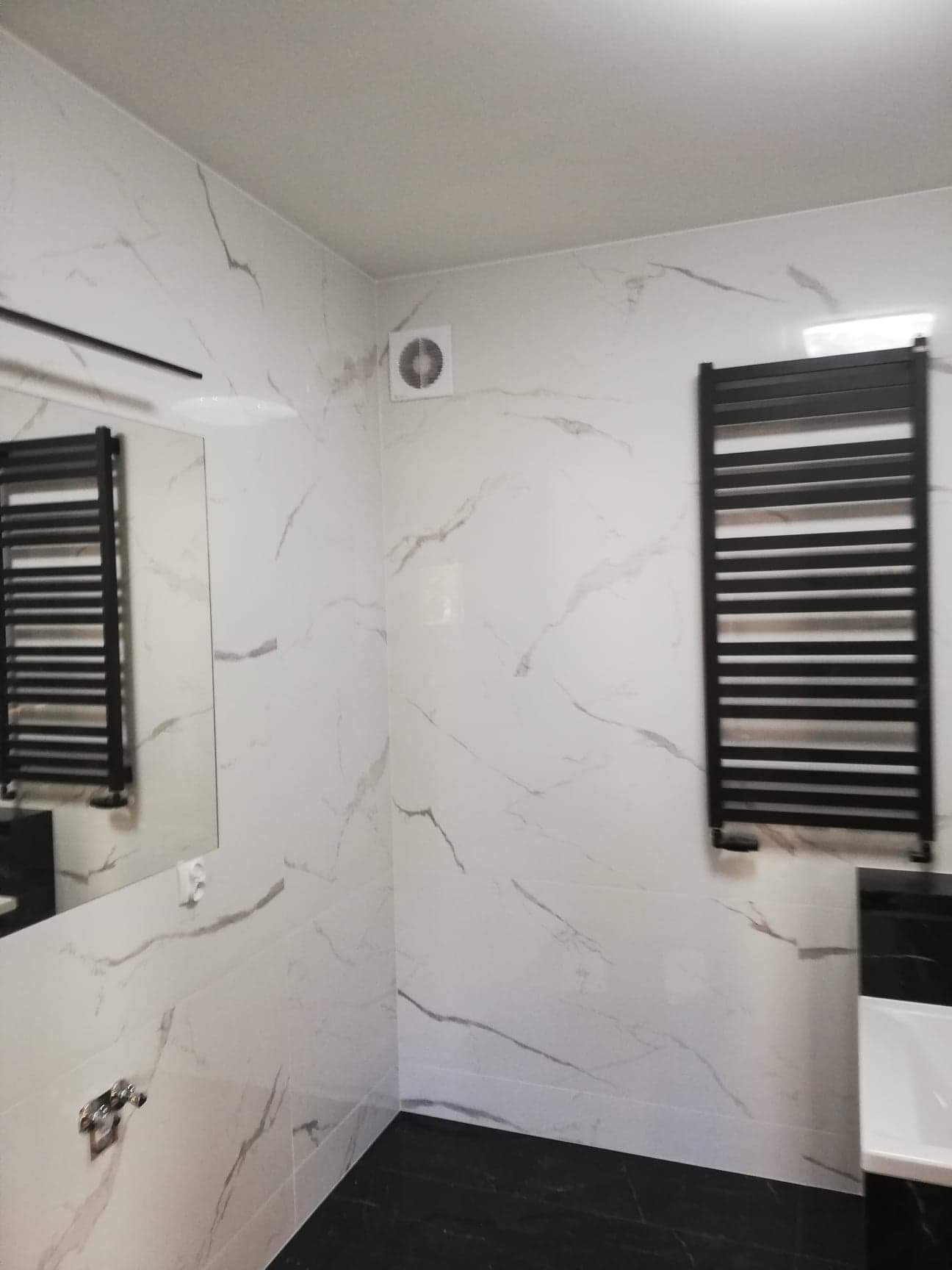 Nowoczesna łazienka z białymi płytkami imitującymi marmur i czarnymi akcentami: grzejnik drabinkowy, podłoga i fragment szafki pod umywalką. Widoczny fragment lustra i wentylator sufitowy.