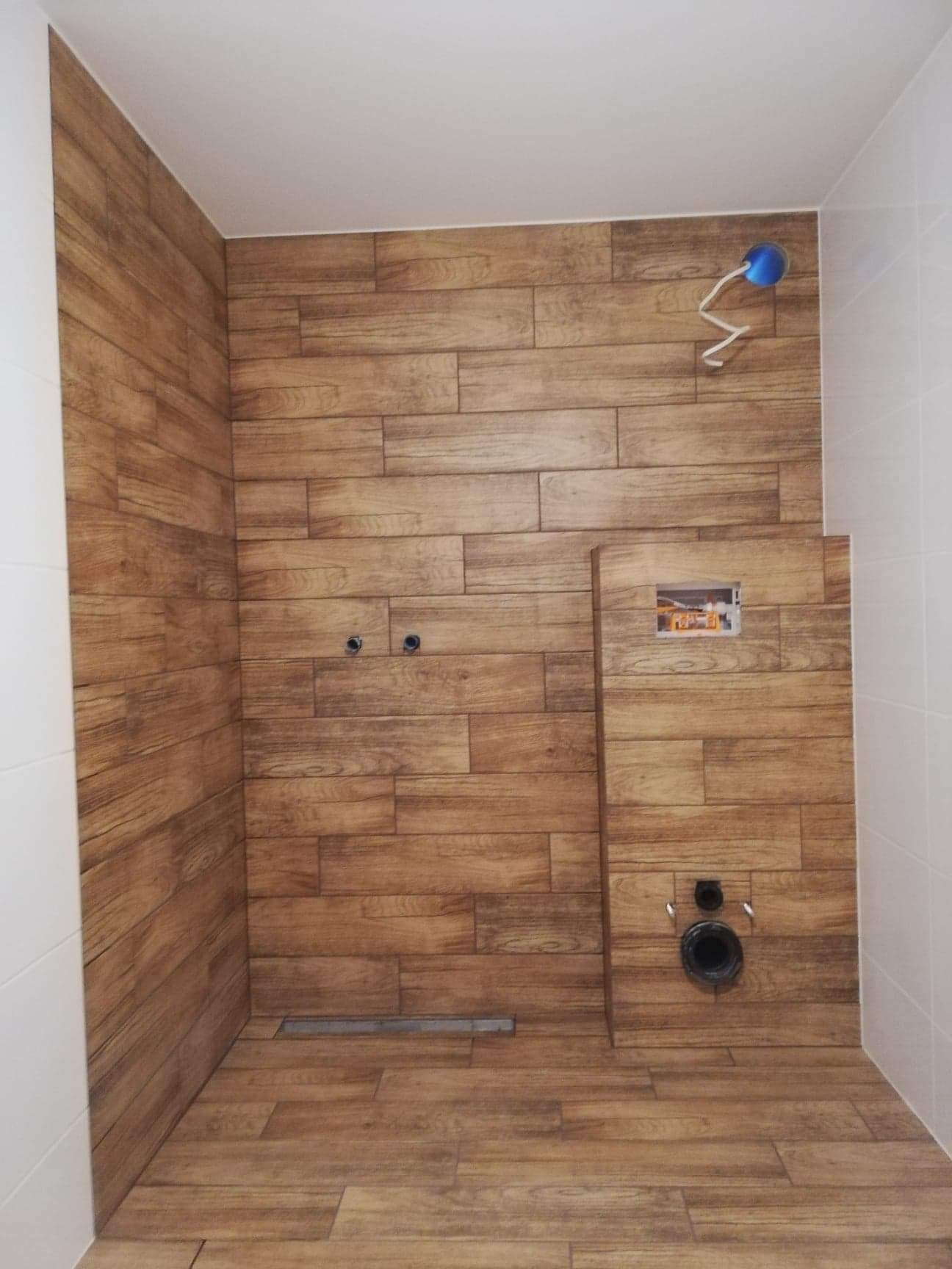 Wnętrze łazienki w trakcie remontu, wyłożone płytkami imitującymi drewno, widoczne otwory na instalacje wodno-kanalizacyjne i odpływ liniowy w podłodze.