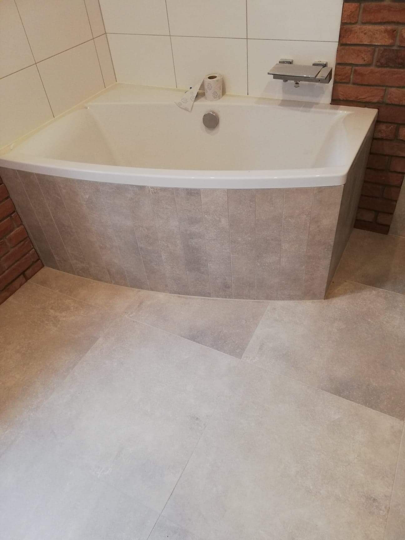 Wykończona łazienka w Dąbcze z narożną wanną obudowaną płytkami imitującymi kamień, jasna podłoga z płytek, fragment ściany z cegły oraz białe płytki ścienne.