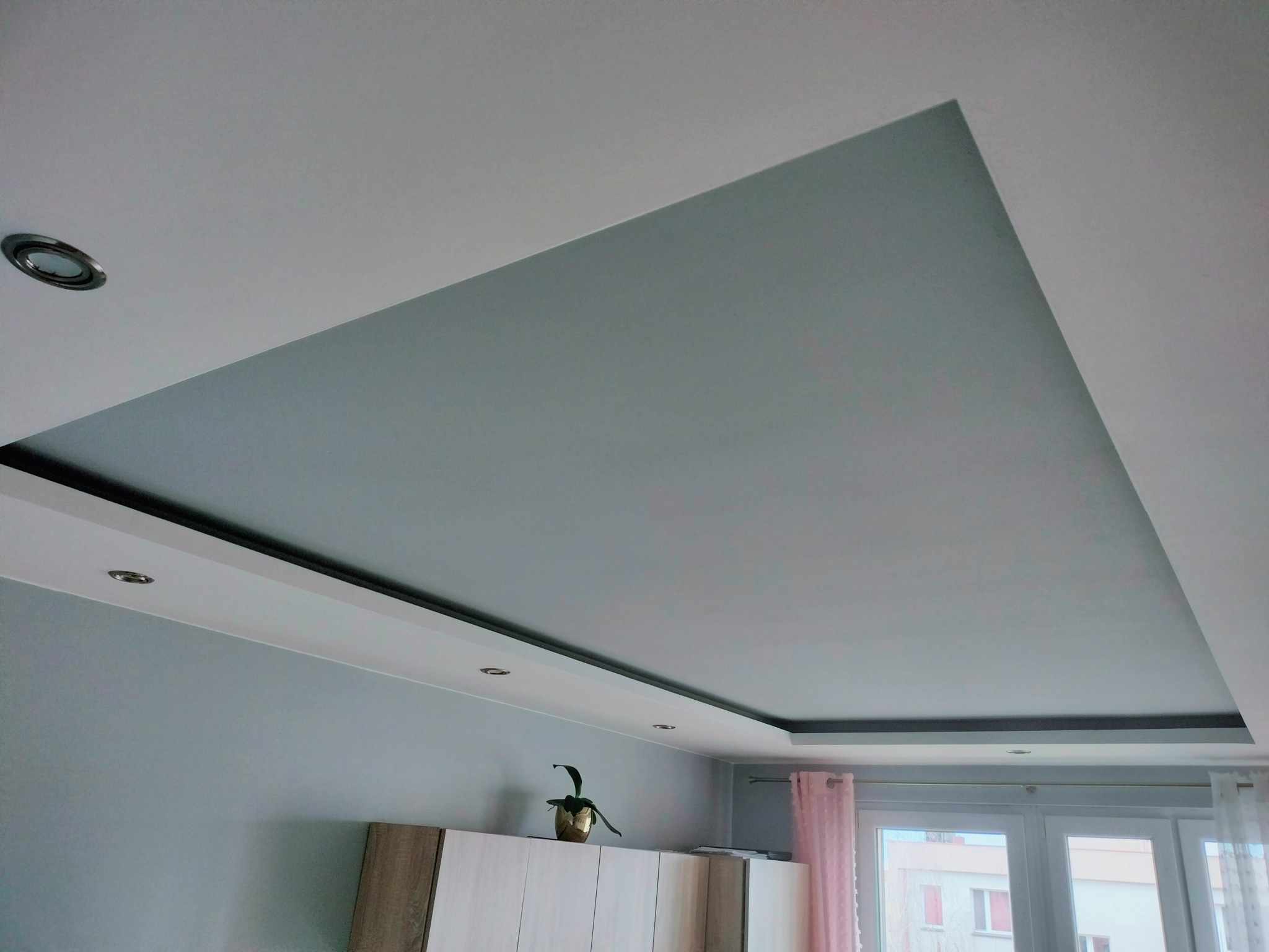 Nowoczesny sufit podwieszany z oświetleniem punktowym i fragmentem ściany w odcieniach szarości, widoczny mebel i roślina doniczkowa na nim.