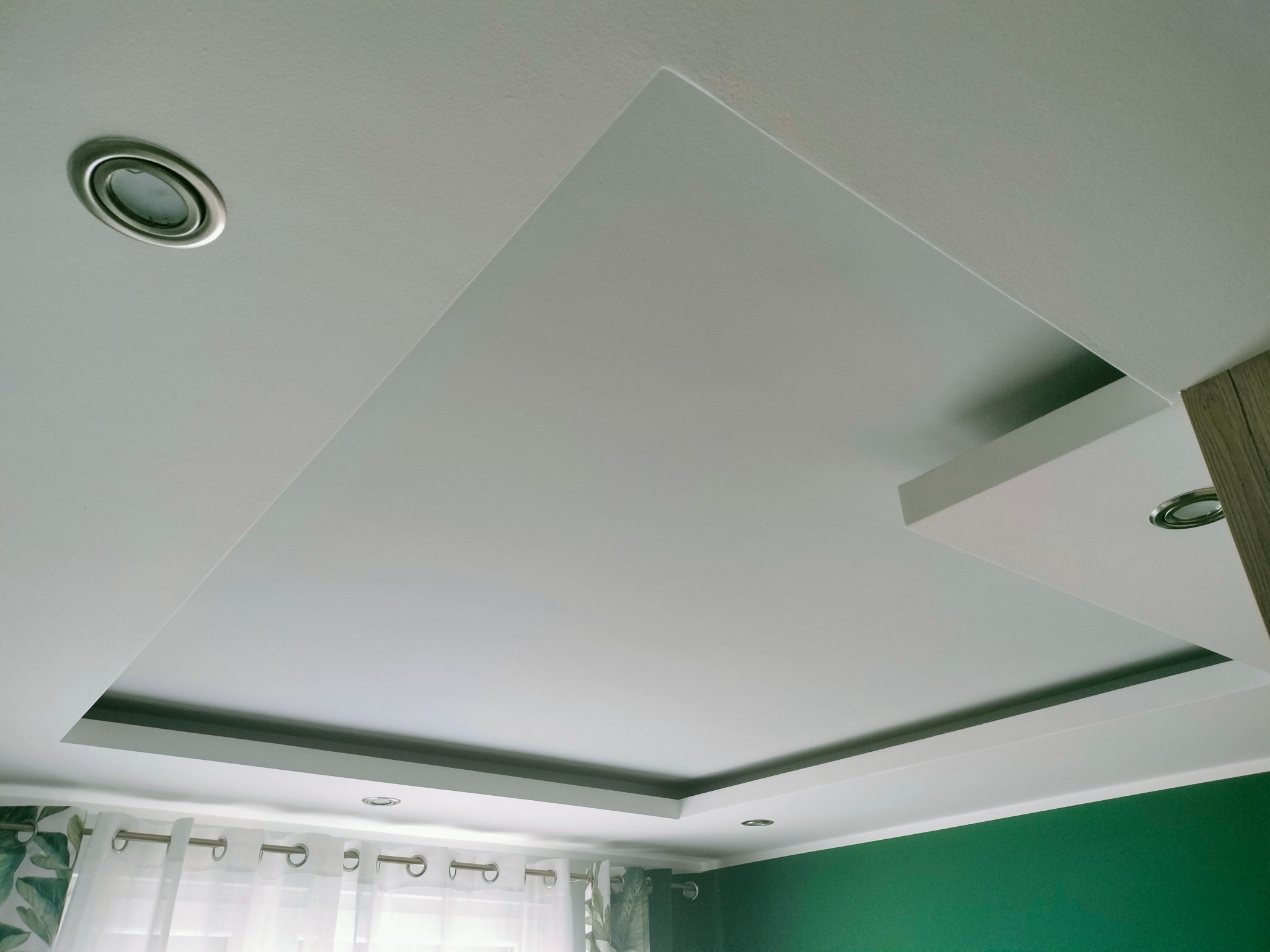 Biały sufit podwieszany z wpuszczonymi punktowymi lampami oświetleniowymi, widok z dołu na fragment pokoju z zieloną ścianą i firankami.