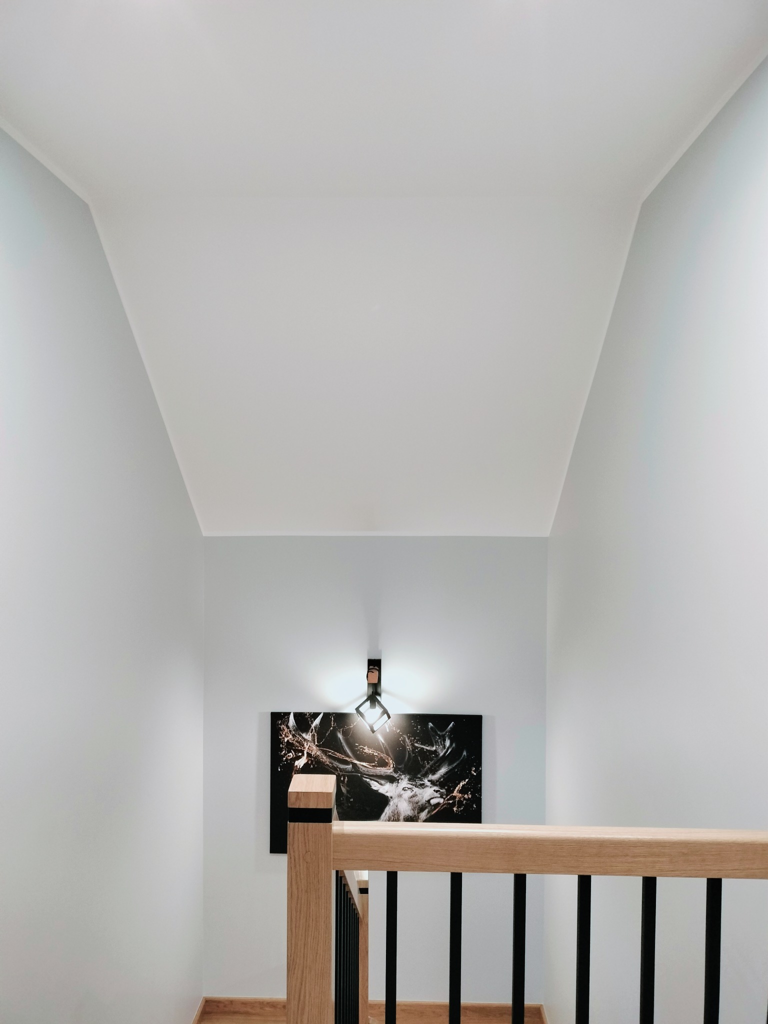 Jasne wnętrze z pomalowanymi na biało ścianami, drewnianą balustradą schodów i obrazem z motywem jelenia podświetlonym kinkietem.