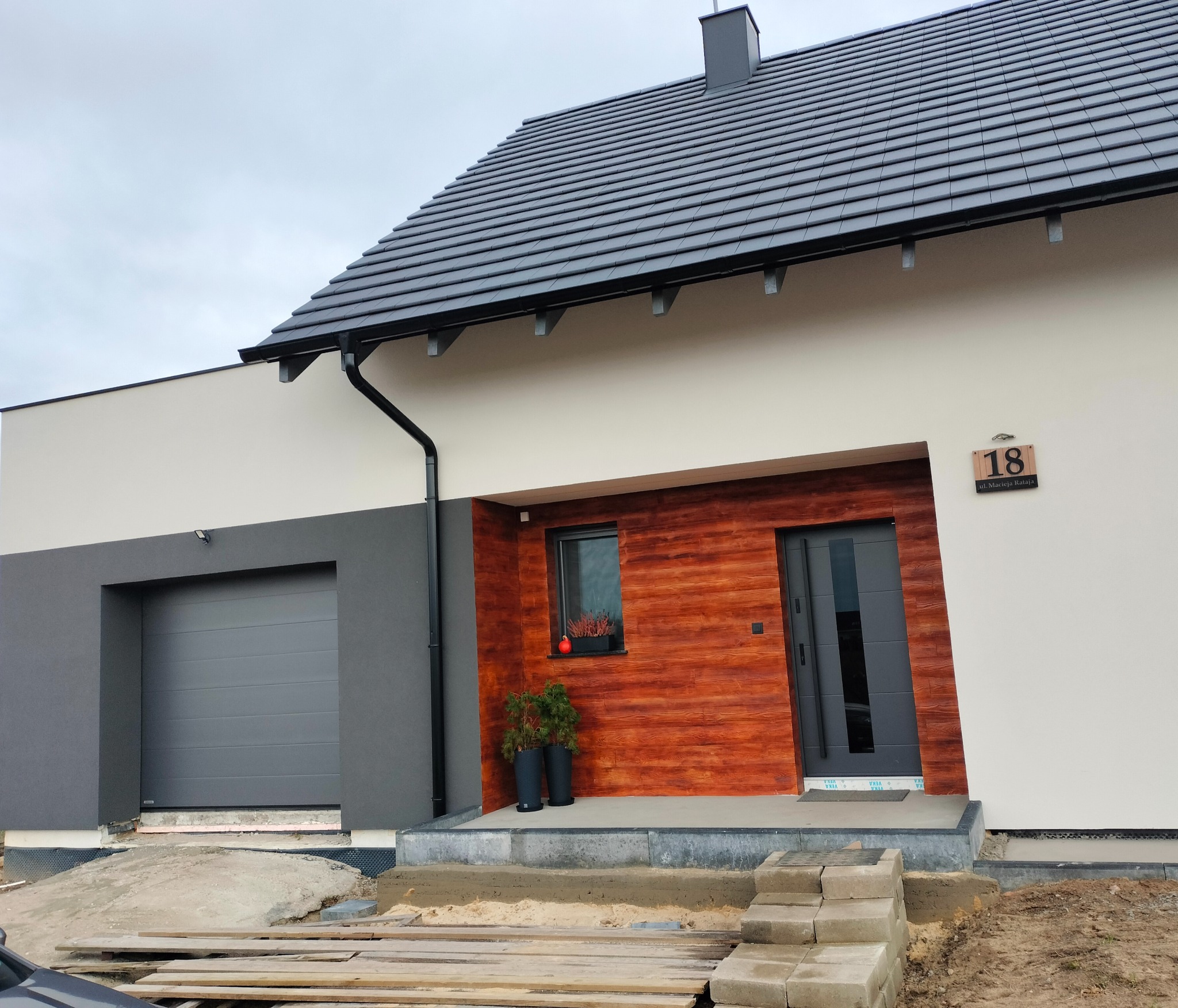 Nowoczesny dom jednorodzinny z elewacją w odcieniach szarości i drewna, widoczny fragment terenu w trakcie prac wykończeniowych.