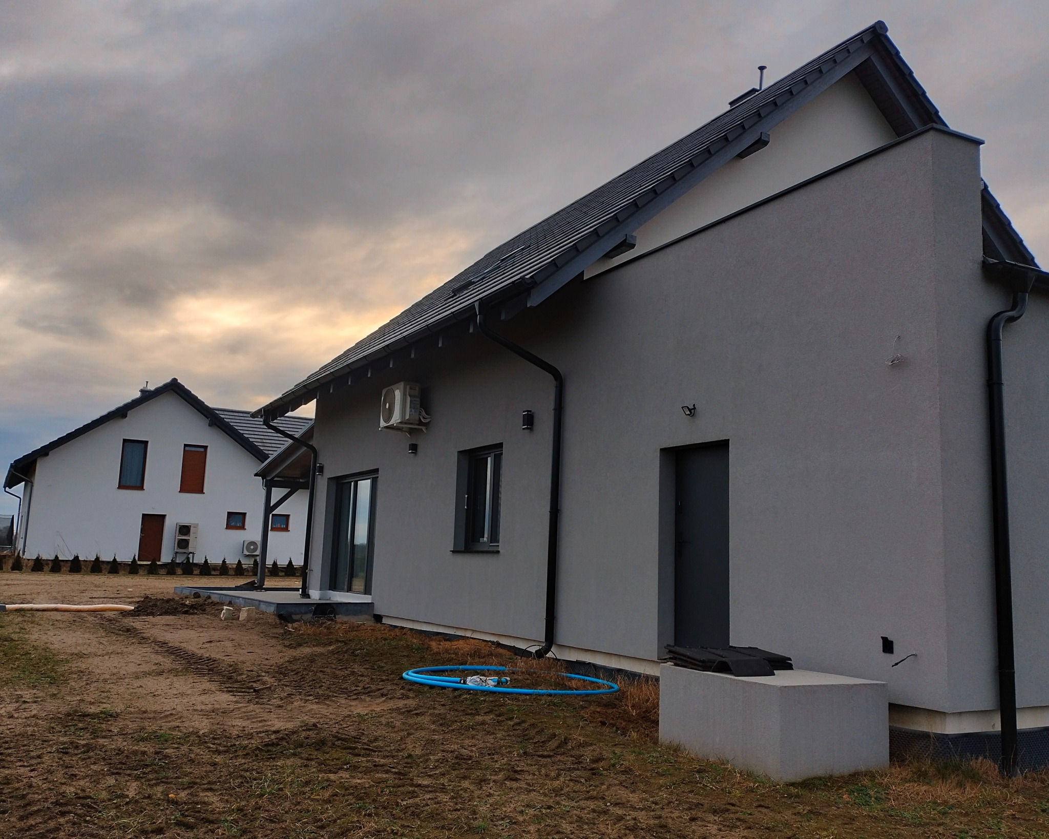 Nowoczesny dom jednorodzinny z szarą elewacją i ciemną stolarką okienną, widok z boku, na pierwszym planie fragment gruntu z niebieskim wężem ogrodowym.