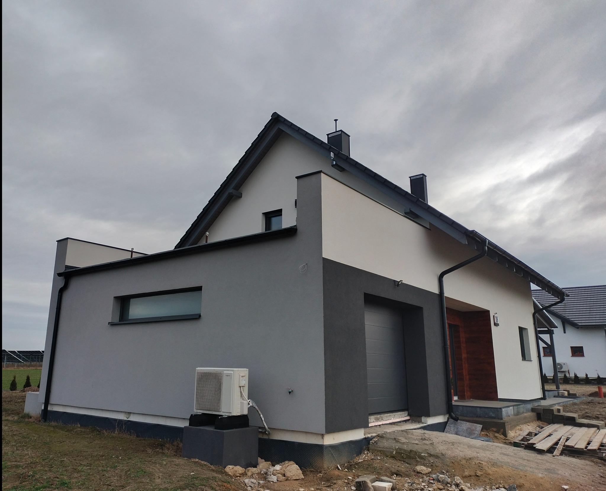 Dwukondygnacyjny dom jednorodzinny z elewacją w dwóch odcieniach szarości i bieli, widoczny klimatyzator na szarej ścianie bocznej, teren wokół domu w trakcie prac wykończeniowych.