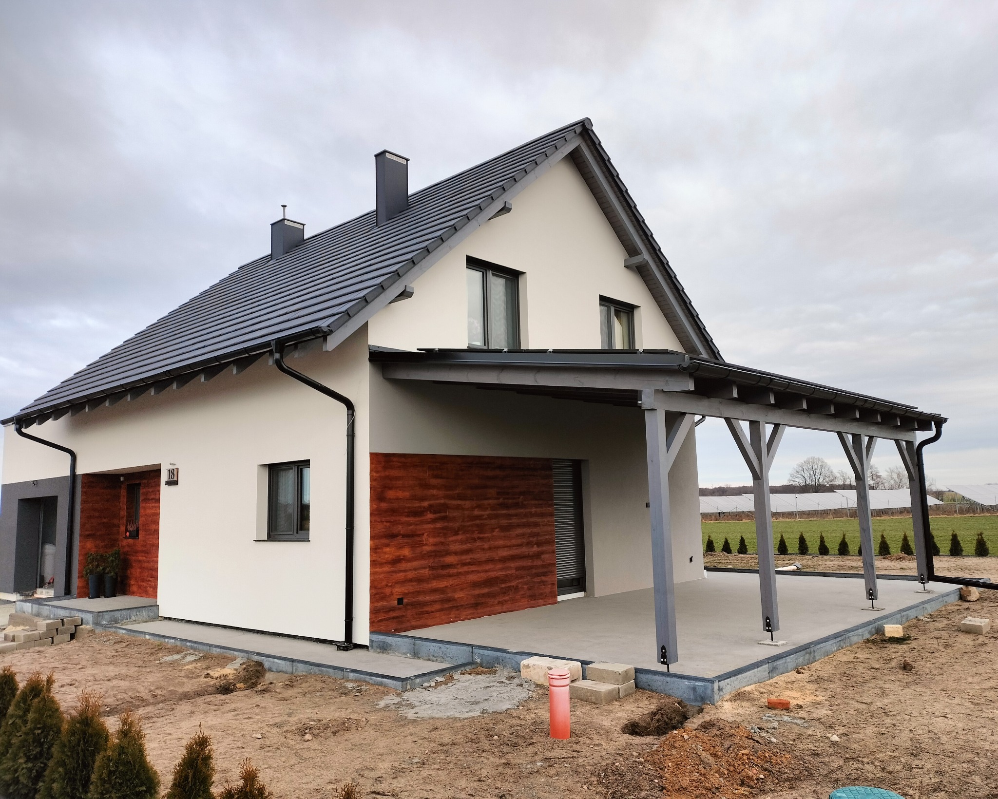 Nowoczesny dom jednorodzinny z białymi ścianami, drewnianą elewacją fragmentu i szarą dachówką, zadaszony taras z drewnianą konstrukcją, widok zewnętrzny w pochmurny dzień.