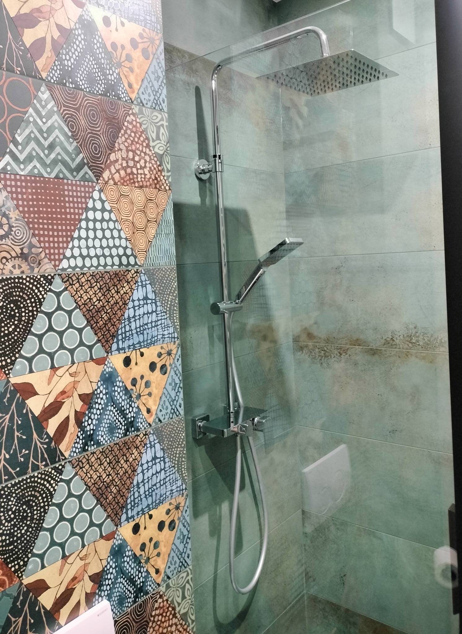 Kabina prysznicowa z deszczownicą, wykończona mozaiką w geometryczne wzory i zielonymi płytkami imitującymi kamień.