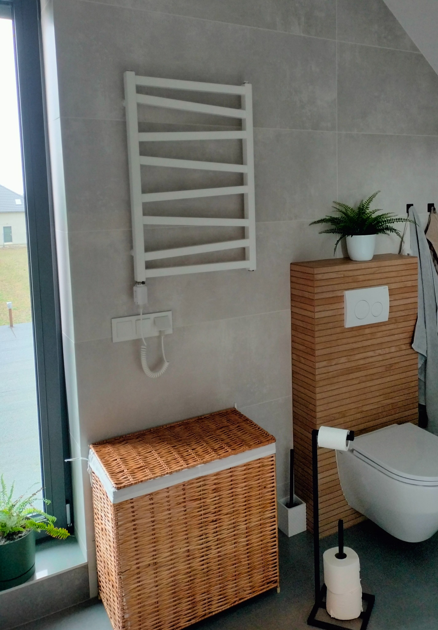 Nowoczesna łazienka z szarymi płytkami, wiszącym grzejnikiem drabinkowym, koszem wiklinowym na pranie, drewnianą obudową toalety i roślinami doniczkowymi.