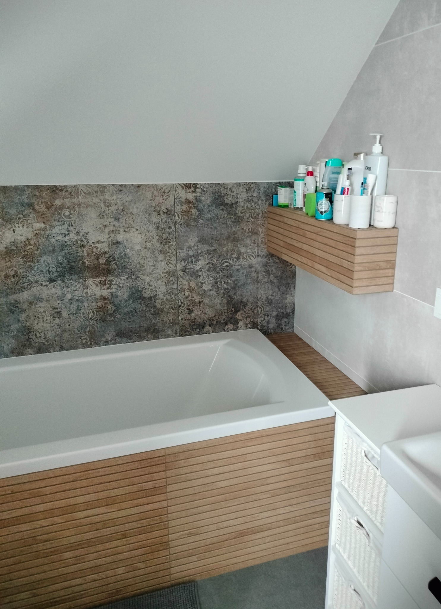 Nowoczesna łazienka na poddaszu z wanną obudowaną drewnianymi panelami, półką z kosmetykami i wzorzystymi płytkami na ścianie.