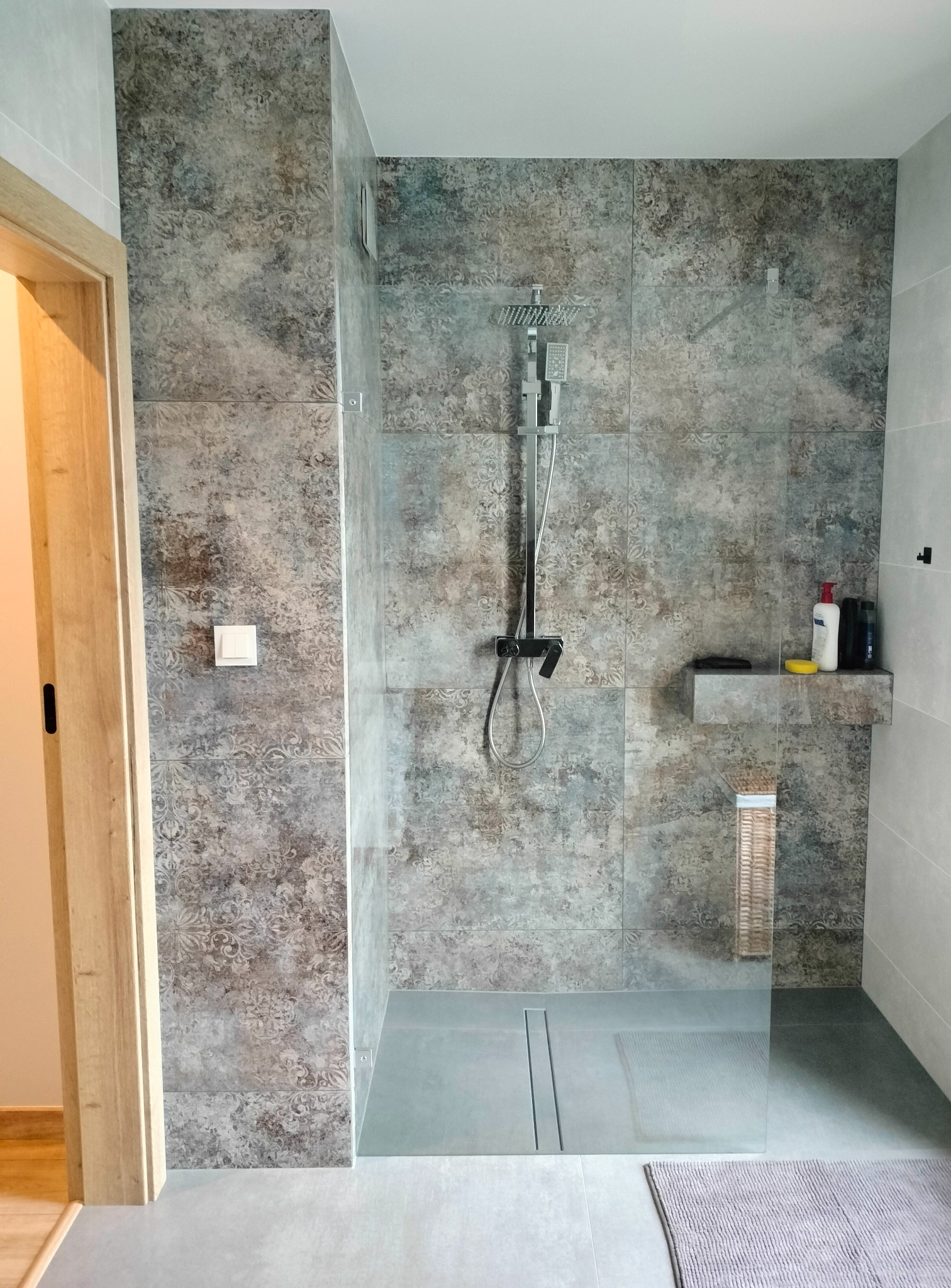 Wykończona łazienka z kabiną prysznicową typu walk-in, wyłożoną płytkami w odcieniach szarości z delikatnym wzorem, z zamontowaną deszczownicą i półką na kosmetyki, widoczny odpływ liniowy...