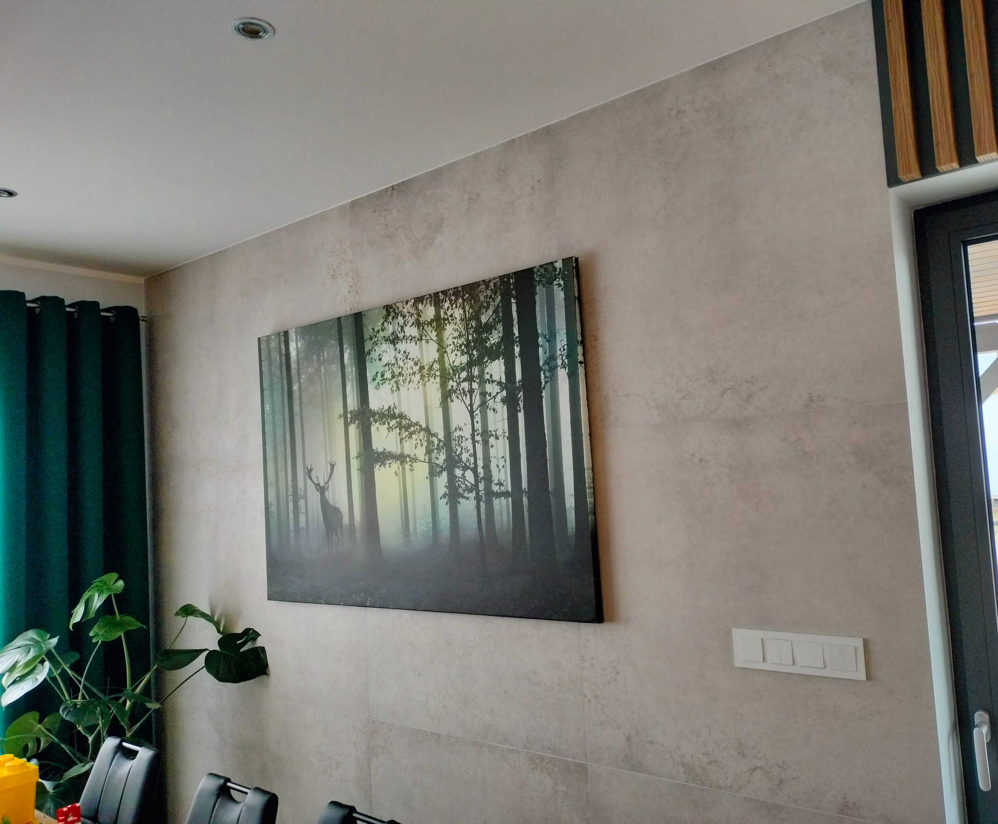 Ściana wykończona płytami imitującymi kamień, z zawieszonym obrazem leśnym z motywem jelenia i włącznikami światła poniżej. Widoczny fragment okna z drewnianymi listwami dekoracyjnymi oraz zielone...