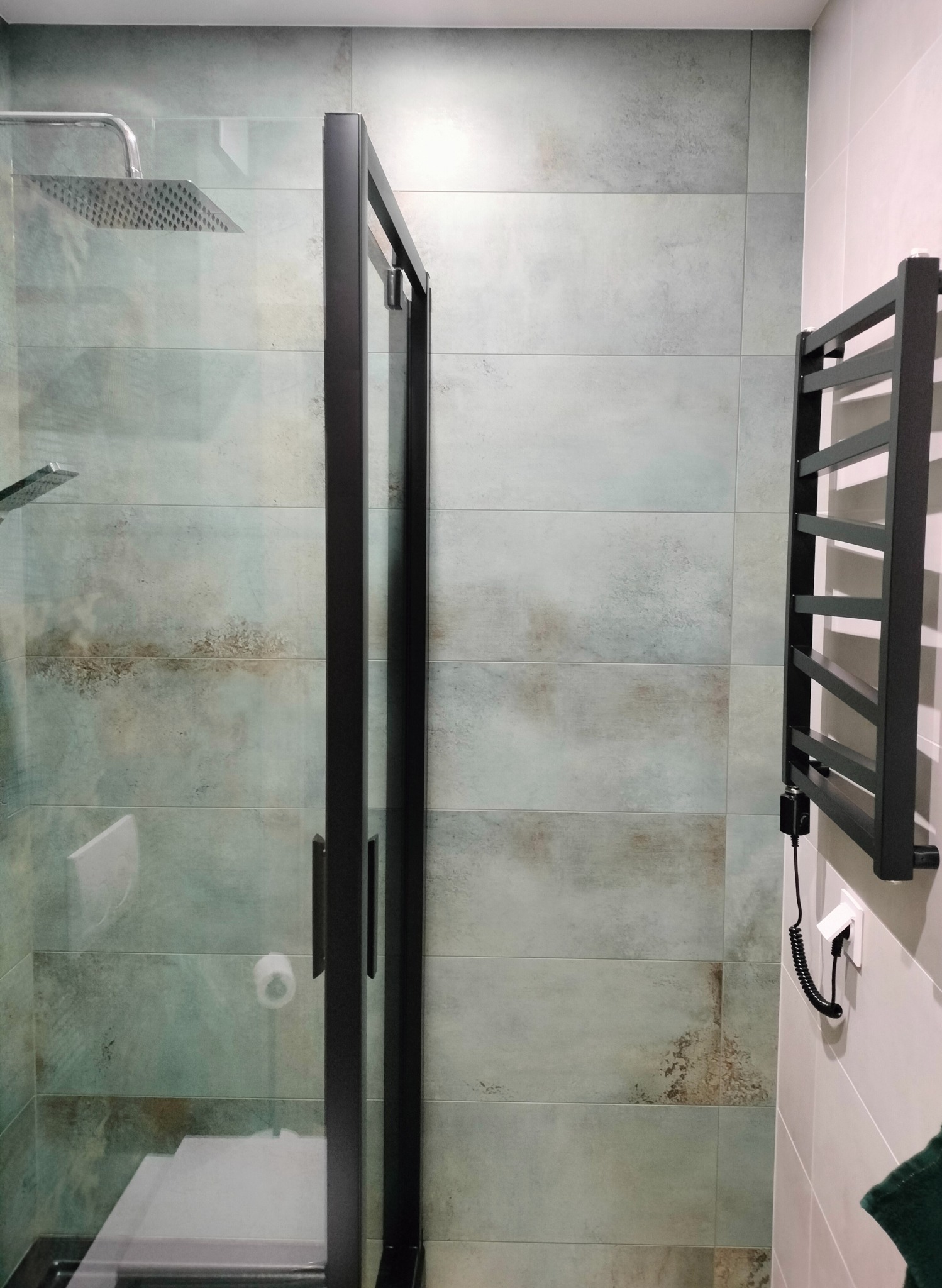 Nowoczesna kabina prysznicowa z czarną ramą i deszczownicą, na ścianach płytki w odcieniach szarości i beżu, obok grzejnik drabinkowy również w kolorze czarnym, z widocznym gniazdkiem elektrycznym...