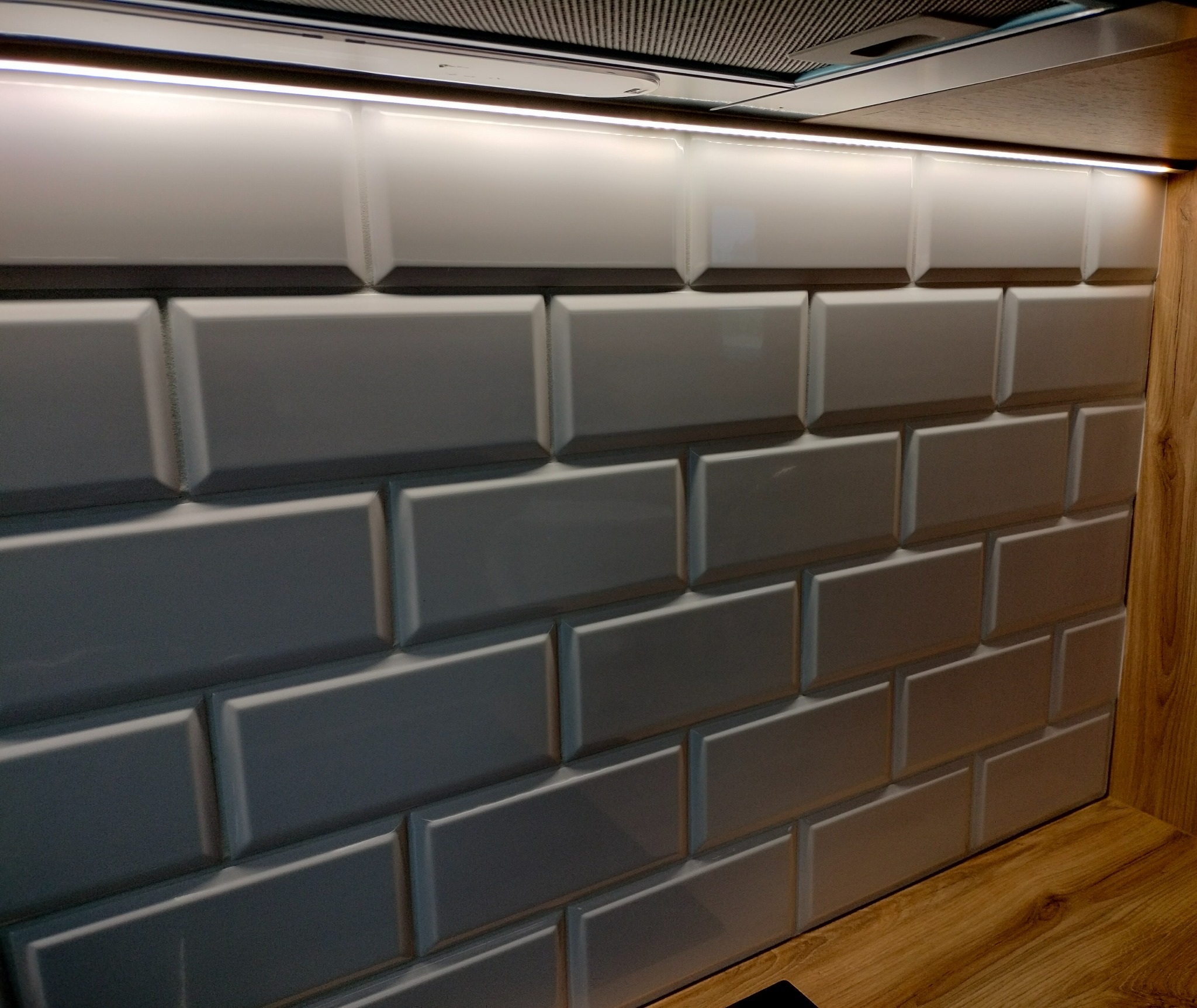 Ściana wyłożona białymi, błyszczącymi kafelkami typu metro w kuchni, oświetlona listwą LED pod szafkami, z widocznym fragmentem drewnianego blatu.