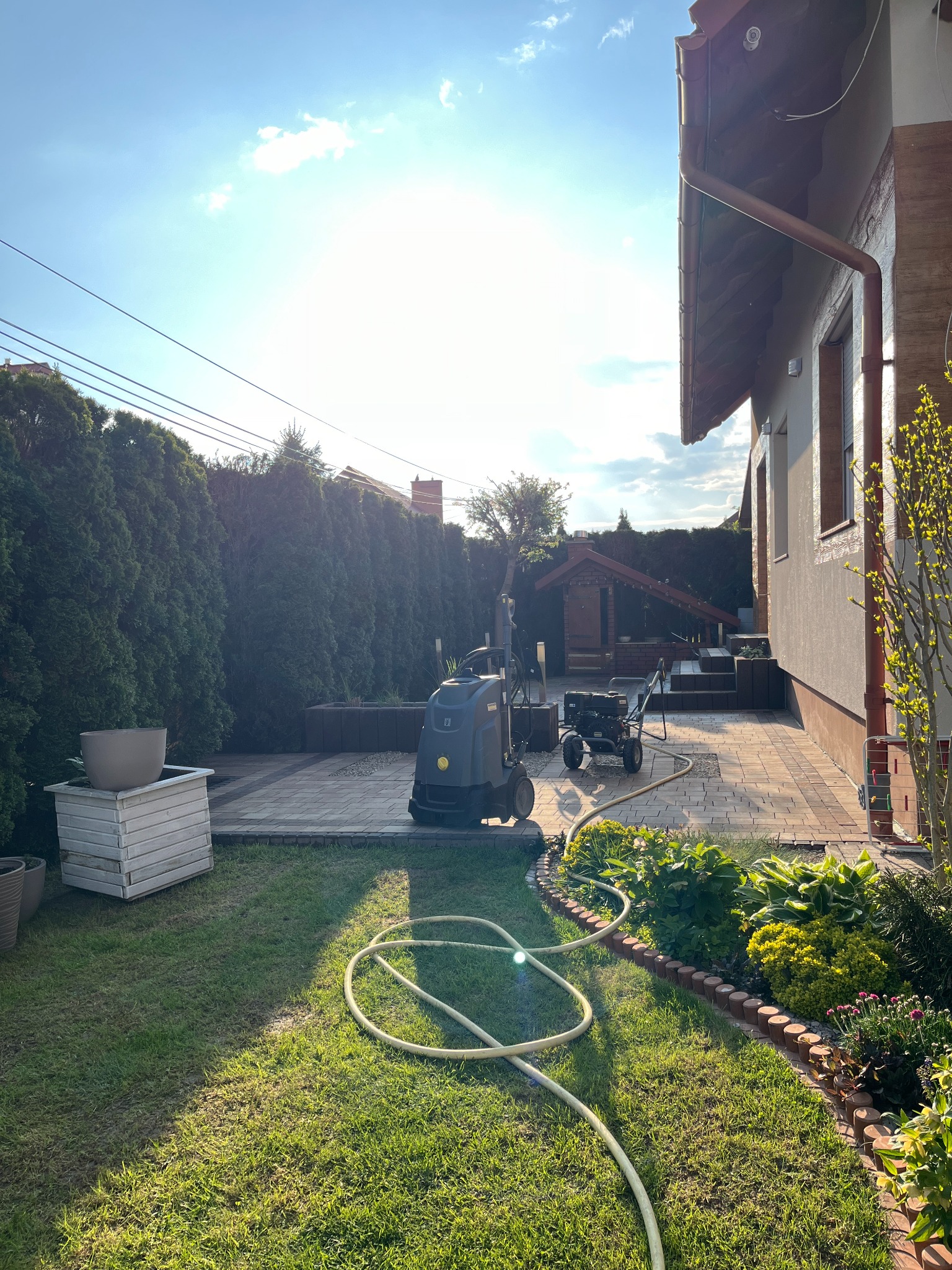 Dwór z kostką brukową, na której stoi myjka ciśnieniowa Karcher oraz wózek transportowy; obok trawnik z wężem ogrodowym, rabata kwiatowa i tuje w tle.