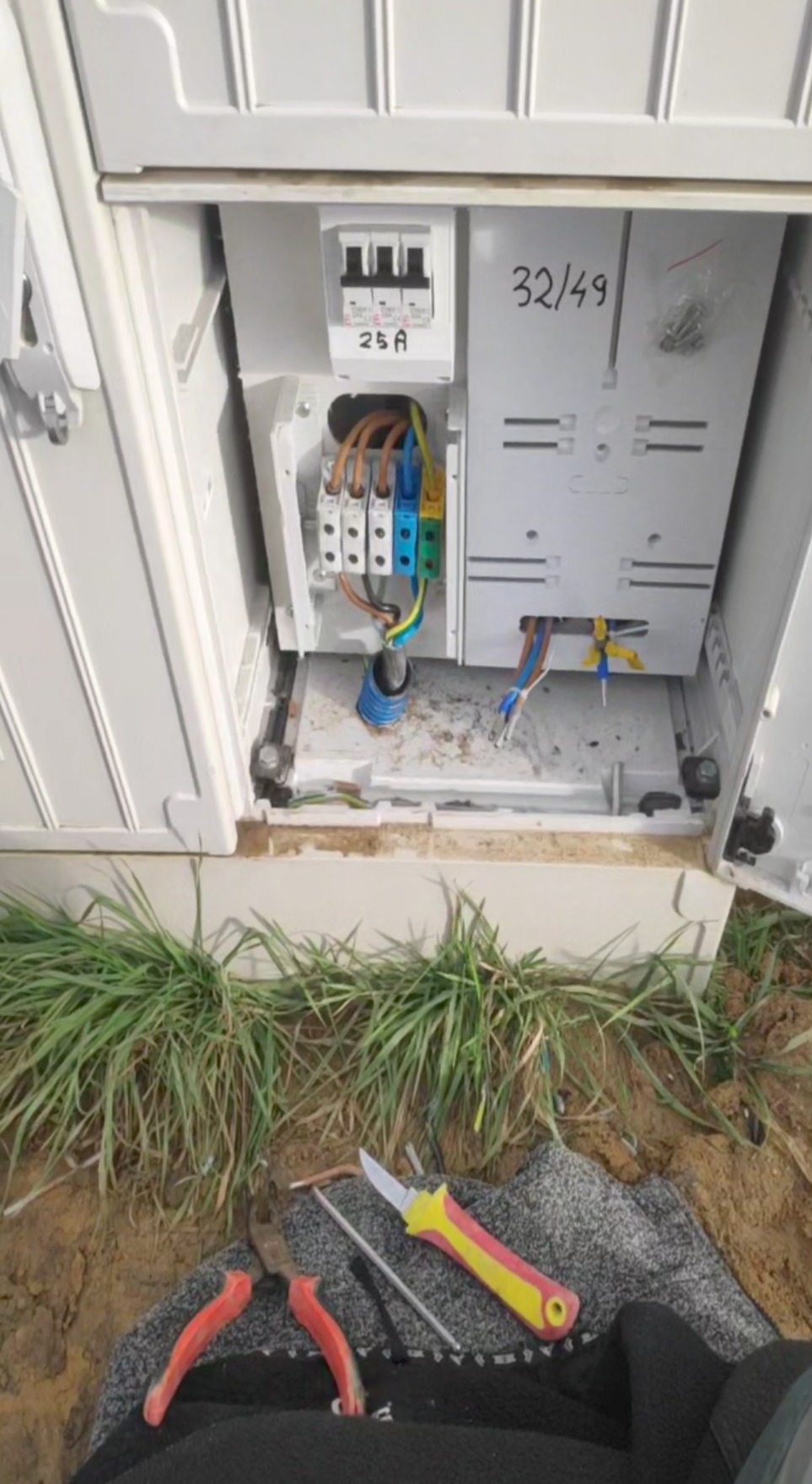 Otwarta skrzynka elektryczna z zainstalowanymi bezpiecznikami i podłączonymi przewodami, widoczne narzędzia: nóż z żółto-czerwoną rękojeścią i kombinerki na ziemi.